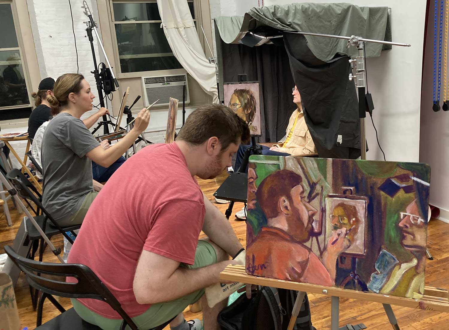 photo of the in progress painting of the painter in context with the painter and model in the background.